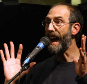 A close up image of Bruce Marcus at a microphone. He is wearing a black t-shirt and glasses, and is gesticulating with his hands.